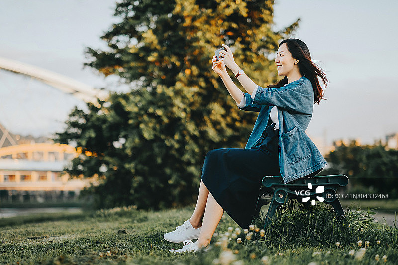 美丽微笑的女人用手机在公园里拍美丽的风景图片素材