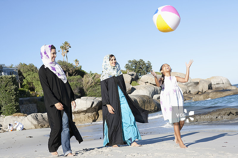 两个阿拉伯妇女看着女孩在沙滩上玩球。图片素材