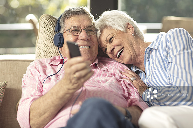 一对幸福的老年夫妇，拿着手机和耳机躺在家里的沙发上图片素材