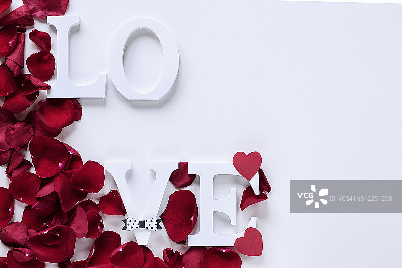 “爱”字的正上方用木制大写字母和小心形纸写着，白色的背景上镶着红玫瑰花瓣。副本的空间。图片素材