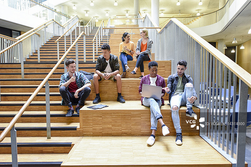 大学里的朋友们坐在现代室内的台阶上聊天，用着笔记本电脑图片素材