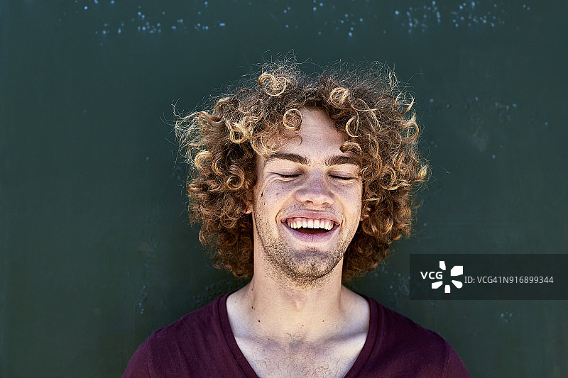 在一堵绿色的墙前，一个留着卷发的笑着的年轻人的肖像图片素材
