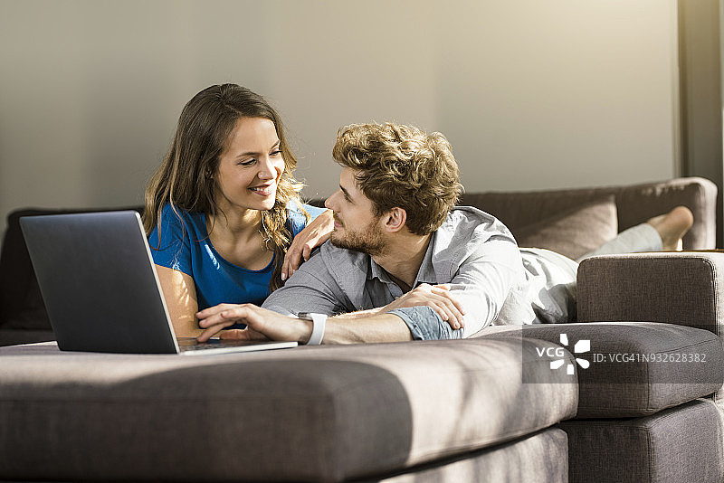 一对微笑的夫妇躺在家里的沙发上，拿着笔记本电脑图片素材