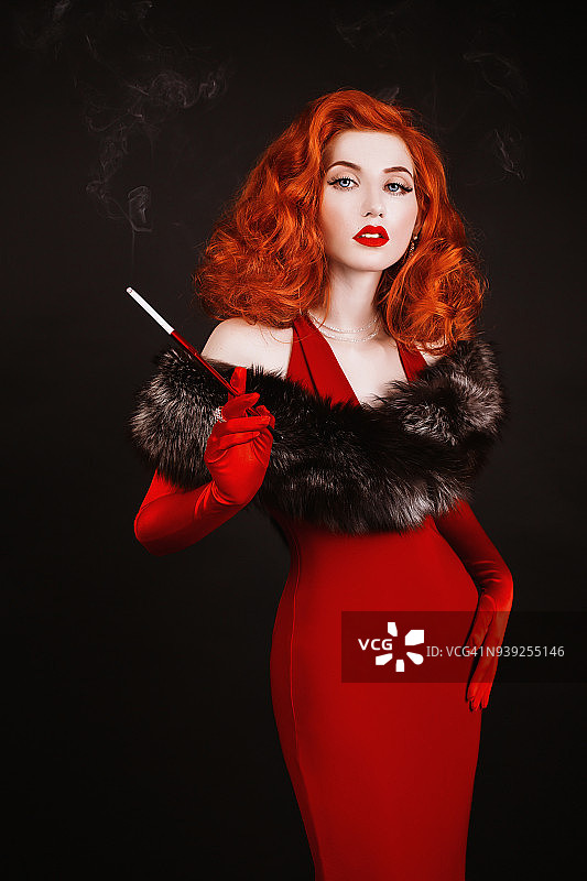 红头发的卷发女人穿着红色的衣服和长手套在黑色的背景下吸烟。女孩穿着复古风格，自然的皮毛和吹口手与白牙齿图片素材