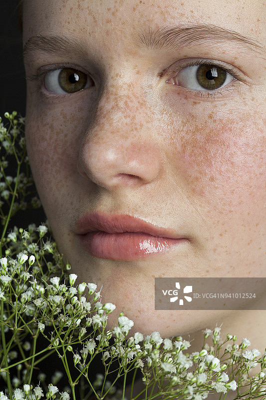 一个十六岁的少女的近距离肖像，带着温柔的微笑，雀斑和苍白的肤色拿着白色的野花。图片素材