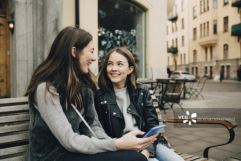 女性朋友微笑着用手机聊天，坐在城市的长椅上图片素材