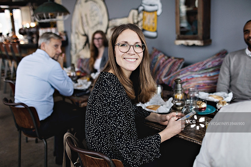 微笑的女人拿着手机的肖像，而坐在桌子上的朋友图片素材
