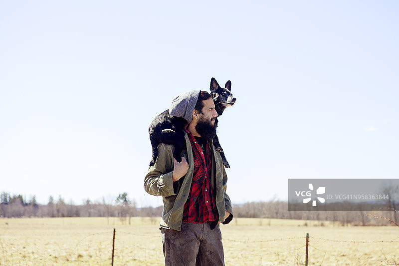 在晴朗的天空下，一个体贴的人背着狗站在田野上图片素材