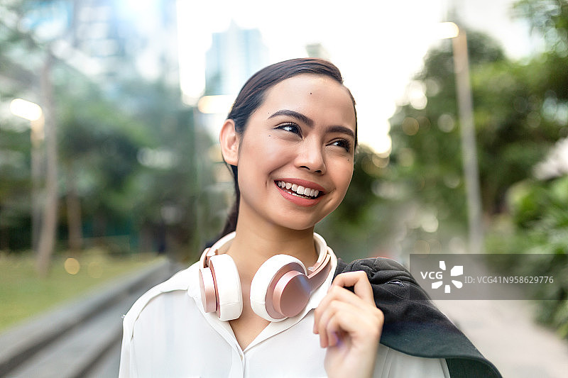 年轻快乐的亚洲女子在公园里散步，肩上扛着一件夹克，脖子上戴着耳机。图片素材
