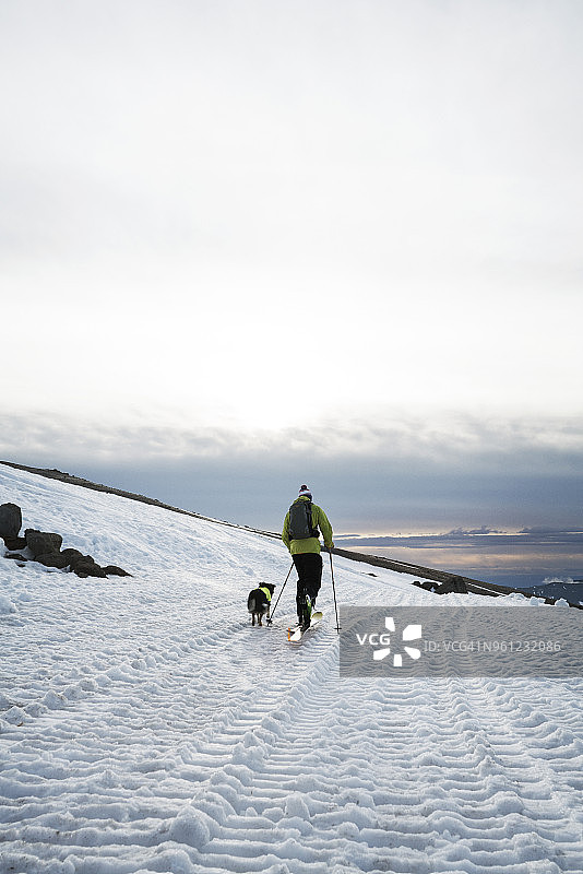 在多云的天空下，一个男人带着狗在白雪覆盖的田野上徒步旅行图片素材