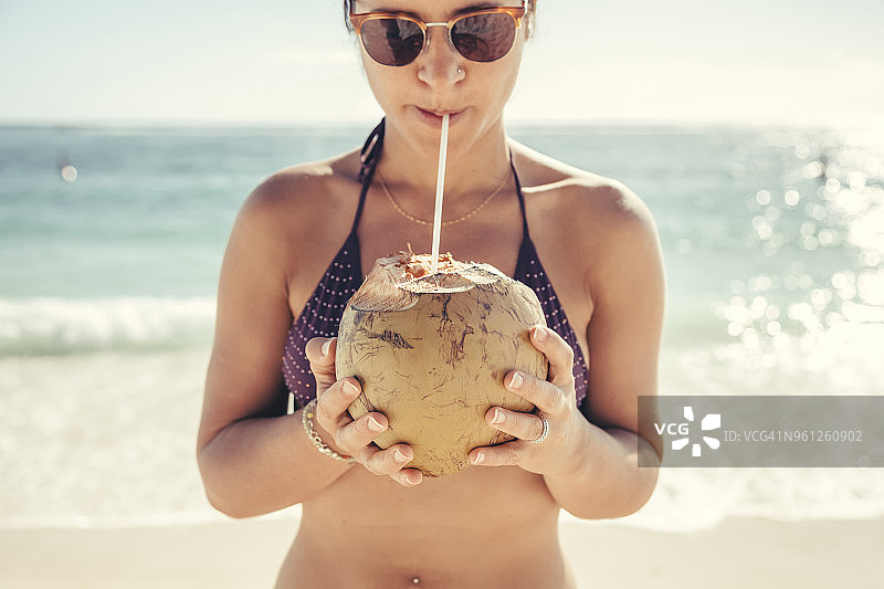 女人戴着太阳镜和比基尼，在海边喝椰子汁图片素材