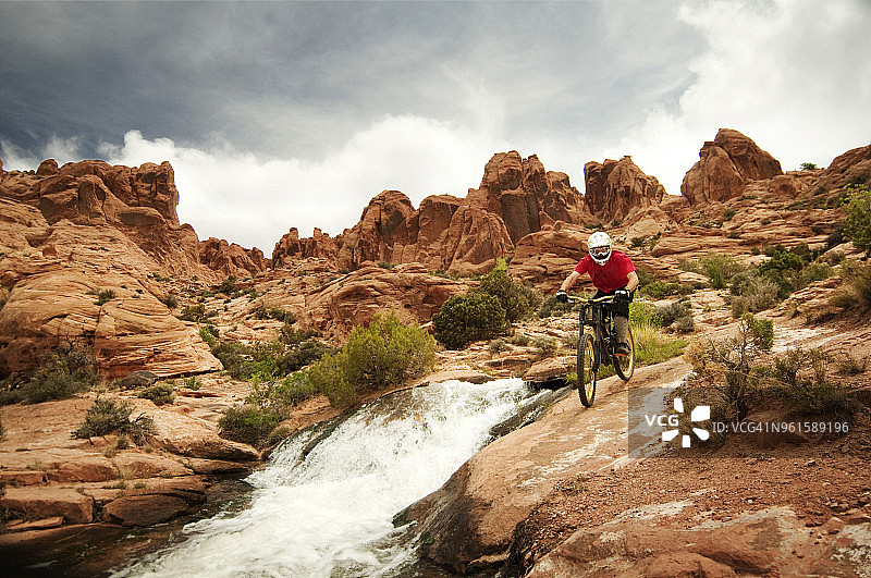 山地自行车手在多云的天空下在河边的岩石上骑车图片素材