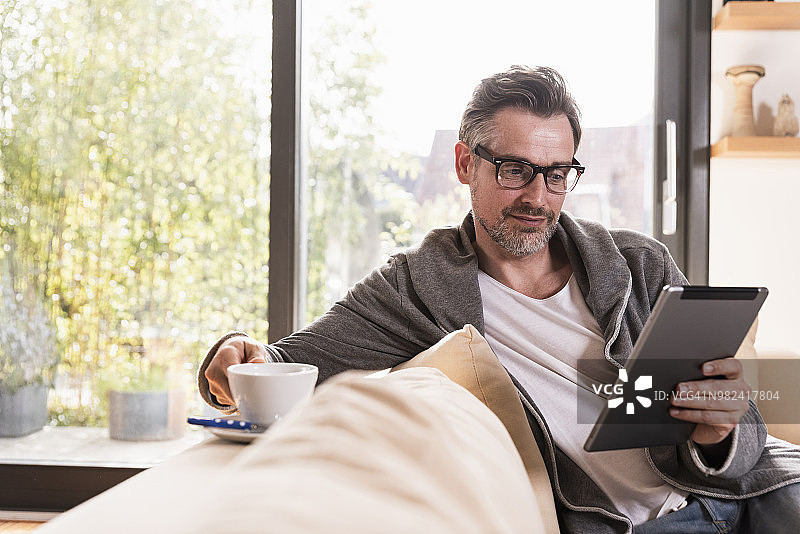 一个成熟的男人拿着一杯咖啡坐在沙发上用平板电脑图片素材
