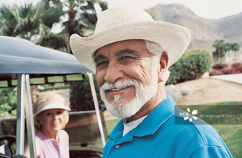 一个年长男子站在高尔夫球场与一个女人在高尔夫球车的背景图片素材