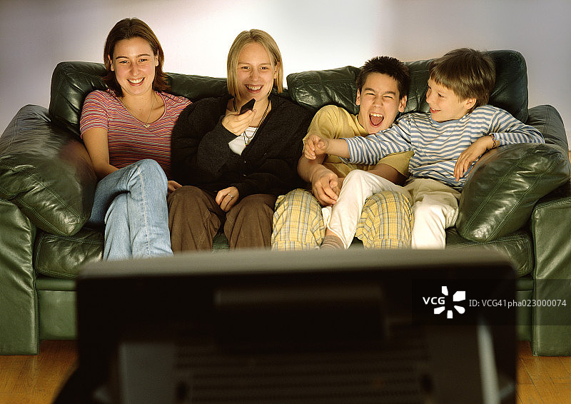 三个年轻人和一个孩子一起坐在绿色的沙发上，笑着，模糊的电视后视图在前景图片素材