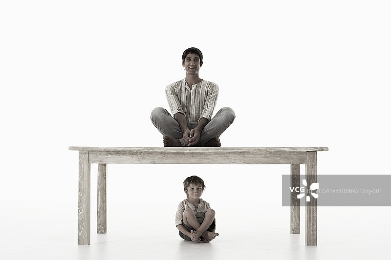 男人坐在桌子上，男孩(4-5岁)坐在下面，对着白色的背景，肖像图片素材