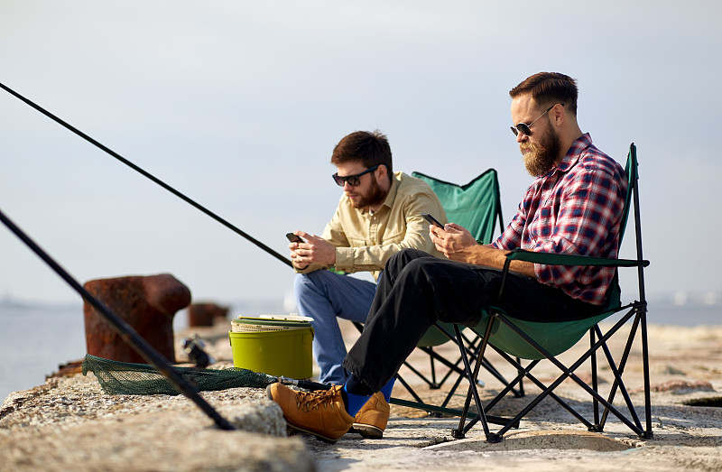 休闲与人的概念——朋友带着智能手机在海边的码头钓鱼。朋友们拿着智能手机在海上的码头上钓鱼图片下载
