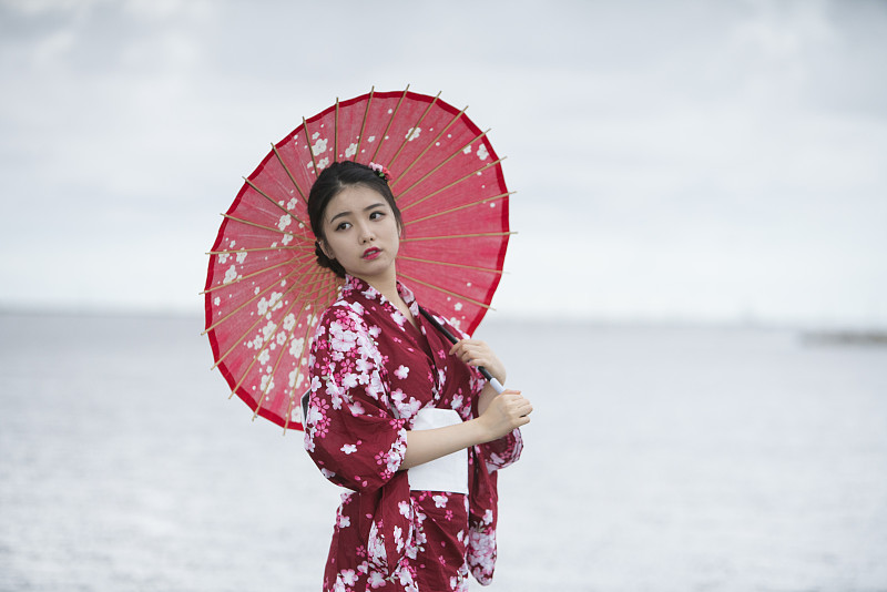 年轻的亚洲妇女的肖像穿着和服与红伞，走在海滩上图片下载