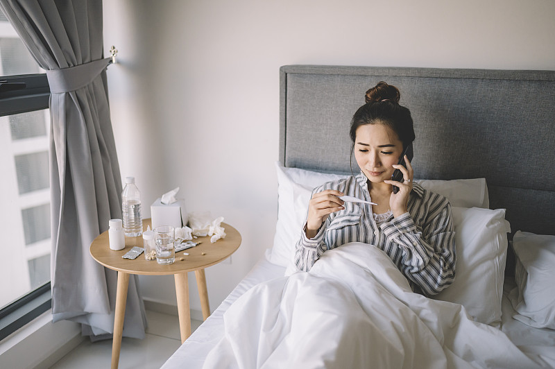 一位亚洲华人女性穿着睡衣在卧室边看床上的温度计边讲电话图片下载