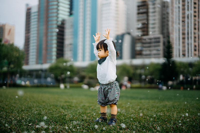 可爱的蹒跚学步的小女孩在城市公园草坪上的绿草上玩肥皂泡图片下载