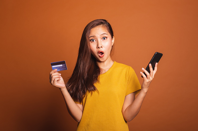 用信用卡和手机站在橙色背景下的震惊女人的肖像图片素材