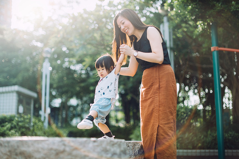 在一个阳光明媚的日子里，快乐的年轻亚洲母亲牵着小女儿的手，扶着她在石头上行走图片下载