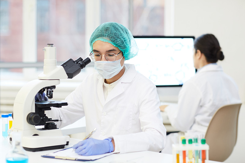 严肃年轻的亚洲实验室研究员，穿着无菌工作服，坐在书桌前，用显微镜记录样品图片下载