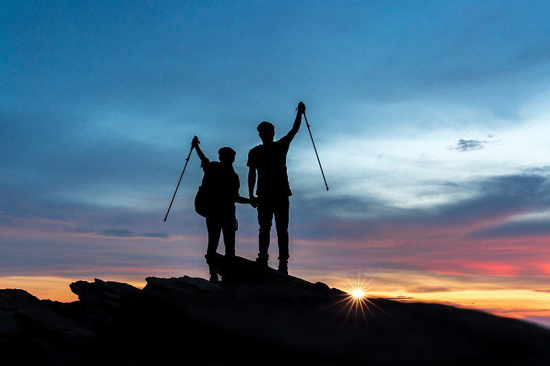 在日落时分，一对年轻的徒步旅行者正在攀登岩石山的顶峰，团队合作，帮助，成功，赢家和领导的概念。图片素材