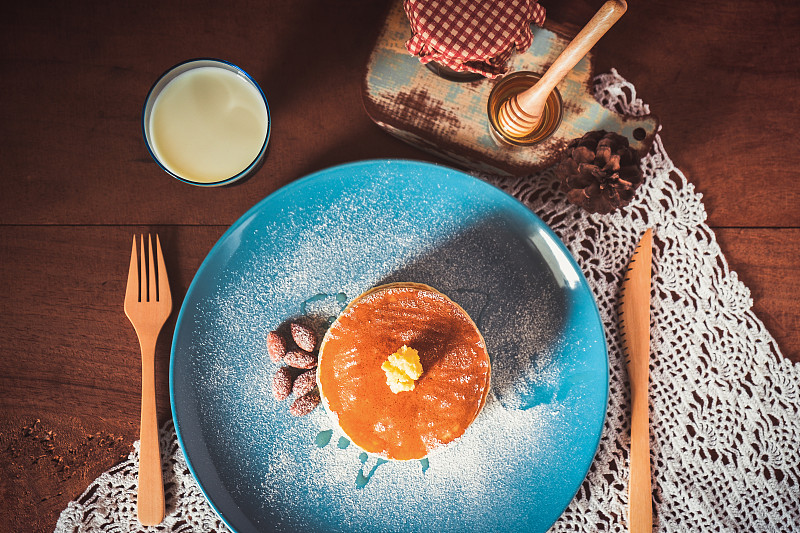 木桌背景上的早餐是蓝色盘子里的牛奶、蜂蜜和杏仁煎饼。图片素材