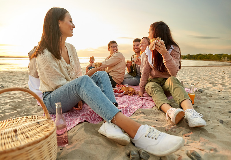 友谊，休闲和快餐概念-一群快乐的朋友吃三明治或汉堡在海滩野餐在夏天。快乐的朋友在海滩上野餐吃三明治图片下载