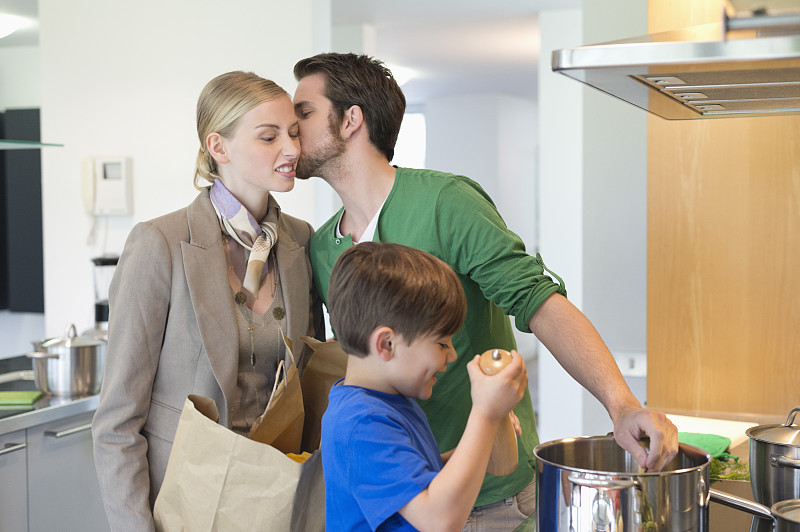 男人亲吻购物归来的妻子，儿子在厨房做饭图片下载