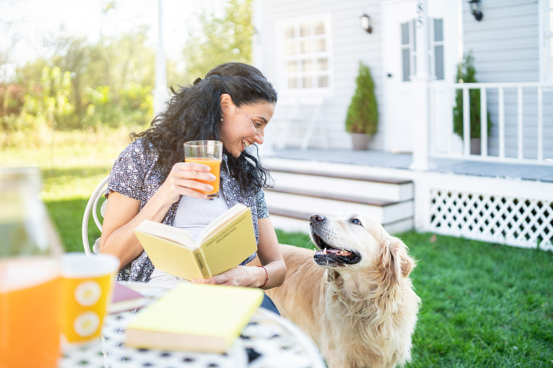 微笑的中年妇女坐在她的金毛猎犬前院里读一本书图片素材