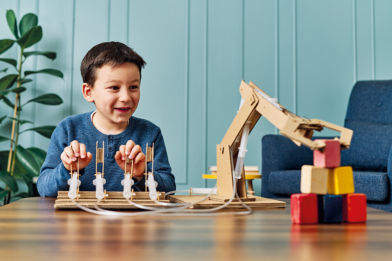 一个6-7岁的孩子发明了一个带纸板和注射器的机械手臂。机器人是他的家庭作业。他在小学时是个成功的学生。图片下载