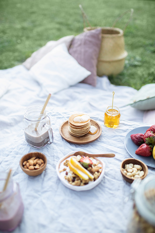夏季健康素食野餐在公园。
水果，可可，蜂蜜煎饼和有机格兰诺拉麦片在亚麻桌布上装饰枕头和柳条篮子。图片素材