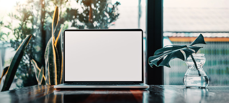 笔记本电脑空白屏幕上的桌子在咖啡馆的背景。笔记本电脑与空白屏幕上的咖啡店模糊背景。图片素材