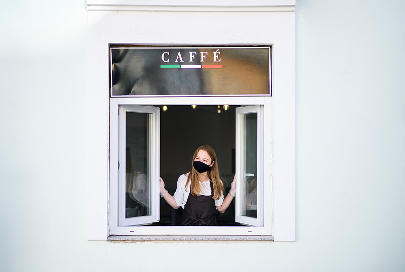 戴着面罩和手套的年轻侍者通过咖啡店窗口端咖啡。图片素材