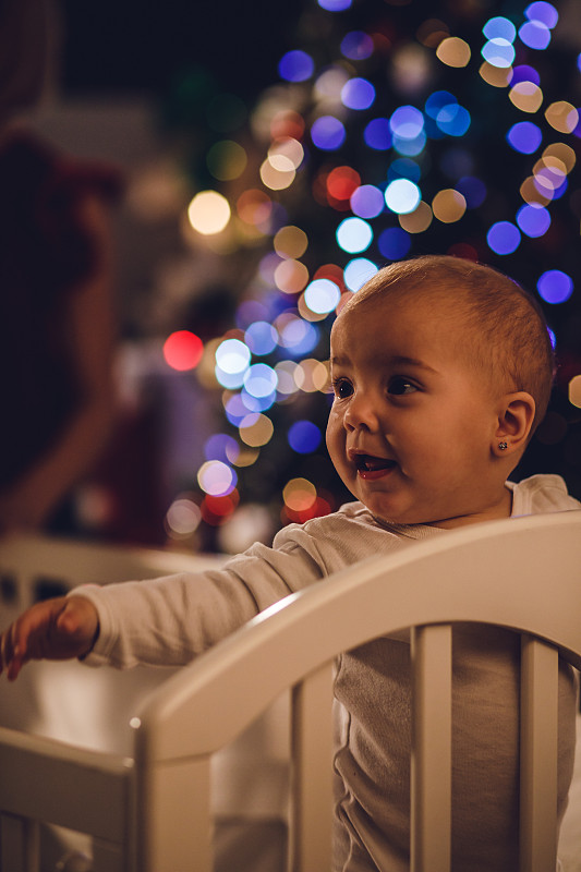 婴儿站在圣诞树旁的婴儿床上图片素材