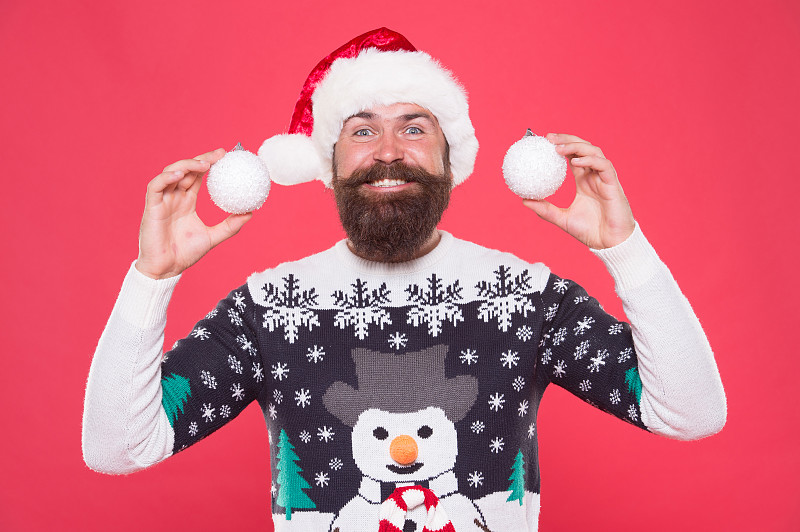 快乐的大胡子小伙穿着暖和的针织毛衣和圣诞老人的帽子，拿着圣诞树装饰雪球庆祝圣诞的冬季假期，新年的装饰图片下载