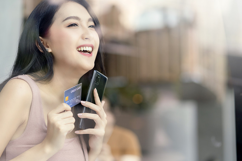 迷人的成年亚洲女性快乐购物网上手提电脑手持信用卡和智能手机购买产品拍摄从窗外的玻璃反射图片下载