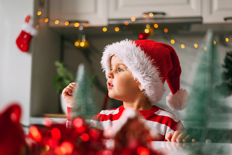 有趣的蹒跚学步的小男孩戴着圣诞帽坐在厨房里，手指着为圣诞节装饰的一边。节日，传统，婴儿食品和健康概念。图片下载