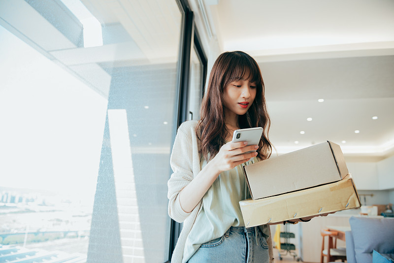美丽的微笑年轻的亚洲女子网上购物与智能手机和接受包裹送货上门服务。图片素材