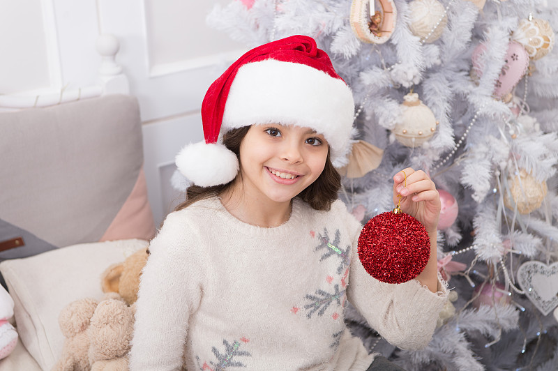 戴着圣诞帽的孩子在装饰圣诞树。华丽的装饰。营造喜庆的氛围。孩子用红球装饰圣诞树。正在装饰圣诞树的小女孩。珍贵的假期活动图片下载