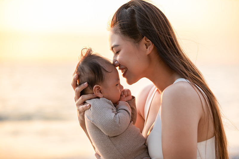 美丽迷人的亚洲妈妈抱着她的新生儿在手和吻在婴儿的头甜美可爱的户外海滩上。快乐的妈妈和小宝宝一起看着夕阳自然的微笑图片下载