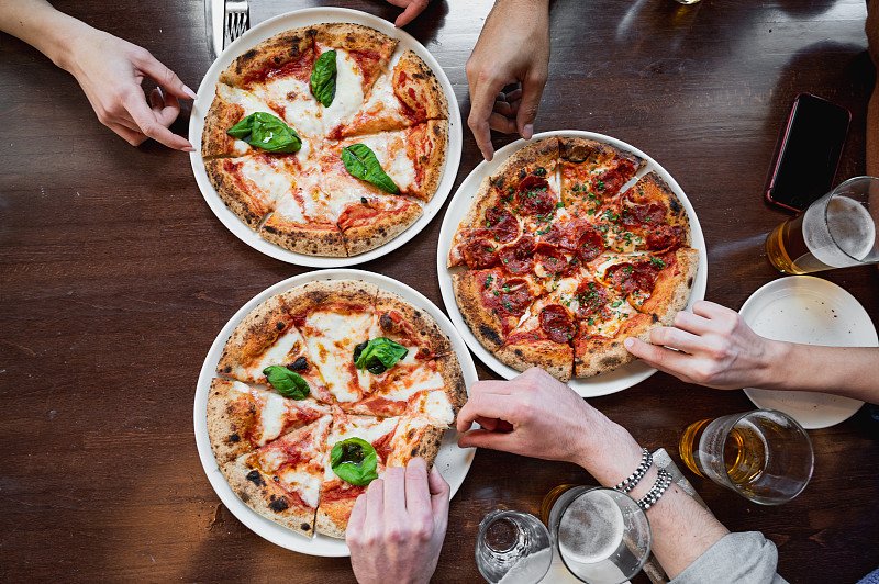 俯视图三个美味的意大利披萨在桌子上。无法辨认的裁剪过的人的手在拿着一片。图片下载