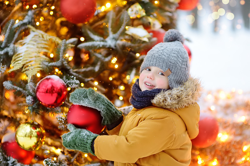 小男孩欣赏装饰着金色、红色球和花朵的人造圣诞树。传统城市户外圣诞市场。城市庆祝装饰图片下载