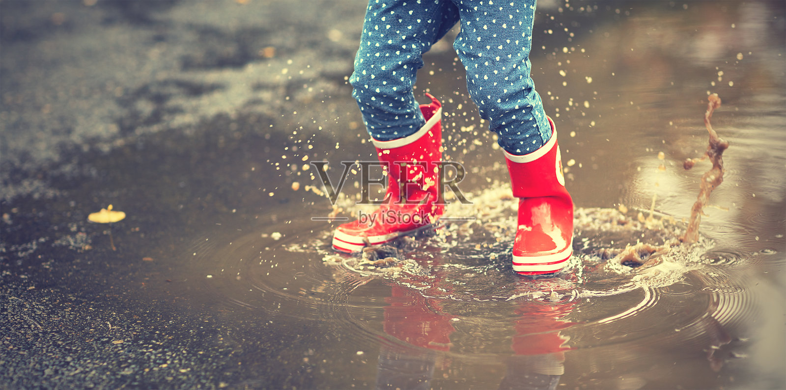 穿着红色橡胶靴子的孩子的腿在水坑里跳照片摄影图片