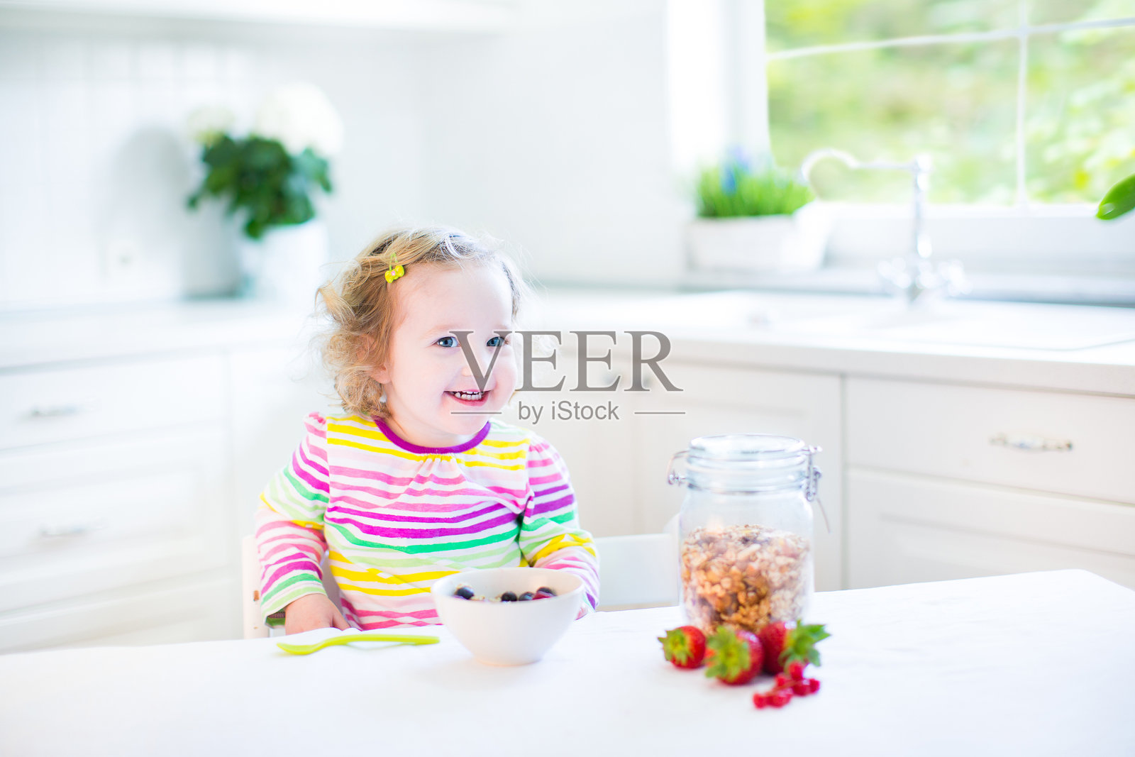美丽的蹒跚学步的小女孩坐在白色阳光明媚的厨房的桌子上照片摄影图片