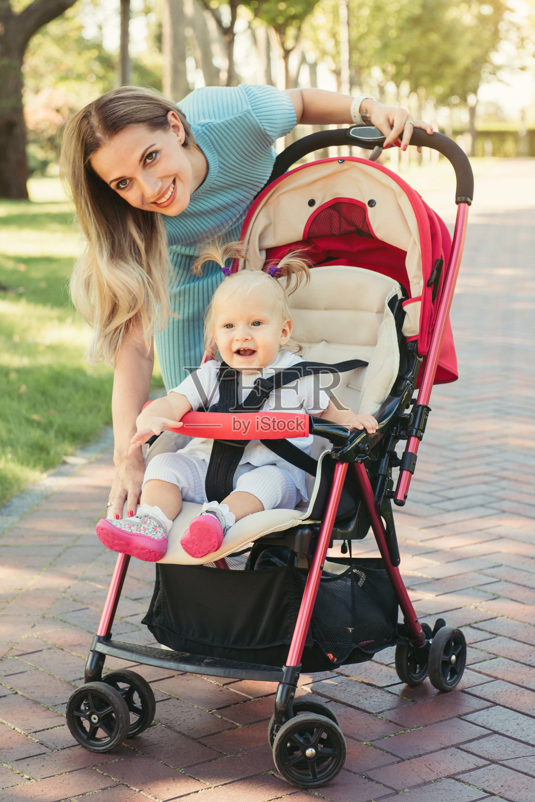 年轻的妈妈和穿着粉色婴儿车微笑的婴儿说话。父母在户外散步的孩子在夏季婴儿车。照片摄影图片