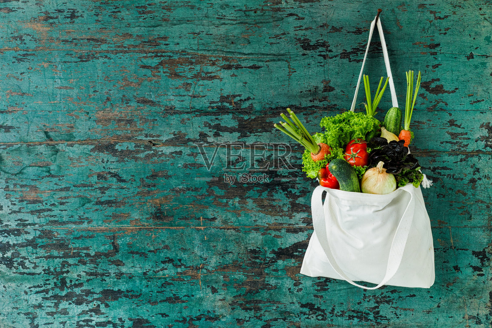 市场新鲜的沙拉蔬菜挂在一个天然棉花可回收的购物袋，用钩子挂在木绿松石色的墙上。照片摄影图片
