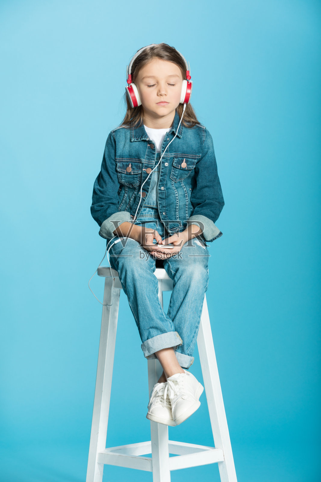 可爱的小女孩戴着耳机使用智能手机，坐在工作室的凳子上播放蓝色照片摄影图片